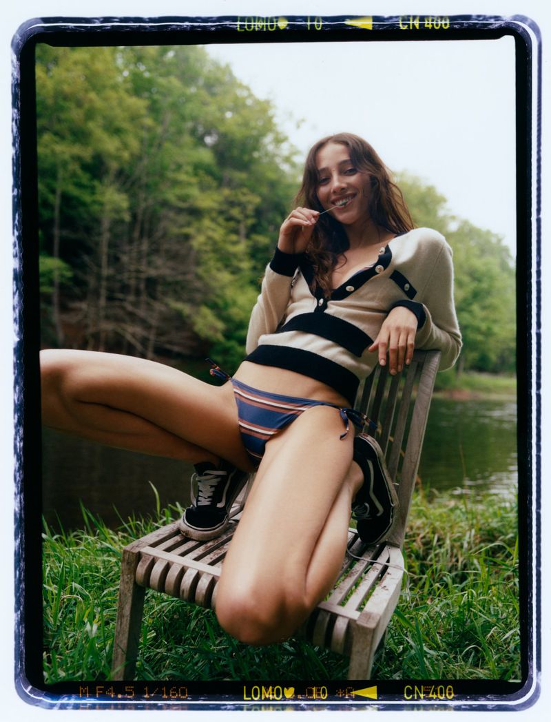 An adult woman is sitting on a wooden chair outdoors near a river, wearing a striped bikini and cardigan, with green trees in the background.