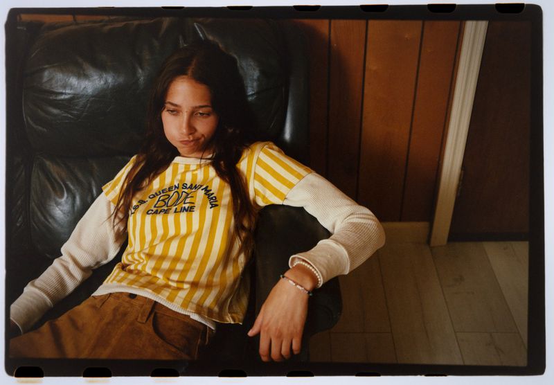 Young adult female relaxing in a black leather armchair at home, wearing a striped yellow shirt with brown pants.