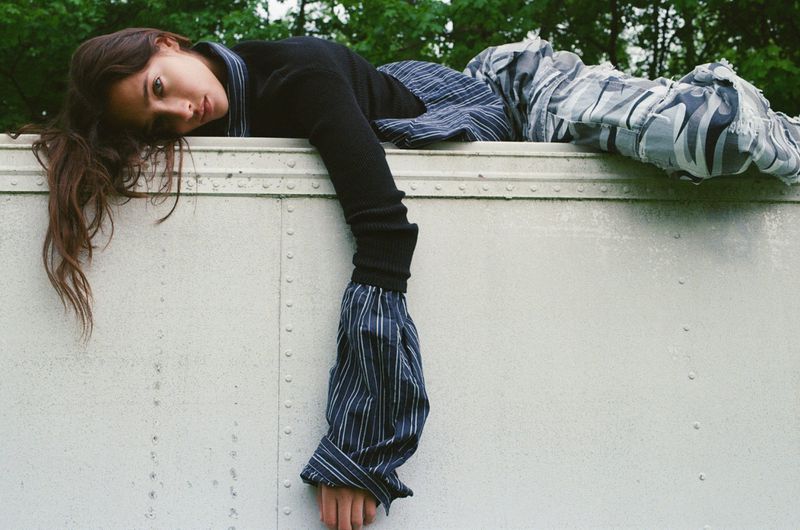 Model Yasmin Wijnaldum lies across the top of a white vehicle wearing a black knit sweater, striped shirt, and patterned pants.