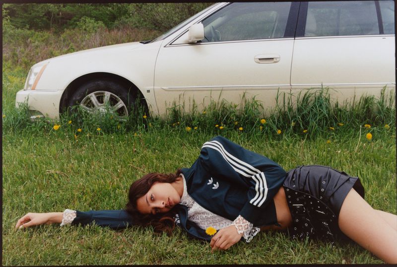 Woman in track jacket poses in a green field near a white car, holding a yellow flower, with soft lighting and a natural, outdoor setting.