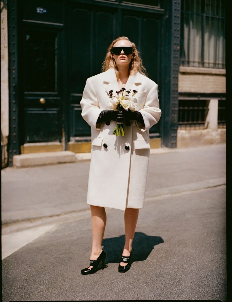 A woman in a cream-colored coat holds a bouquet of flowers on a city street, wearing sunglasses and black gloves for a fashion editorial.