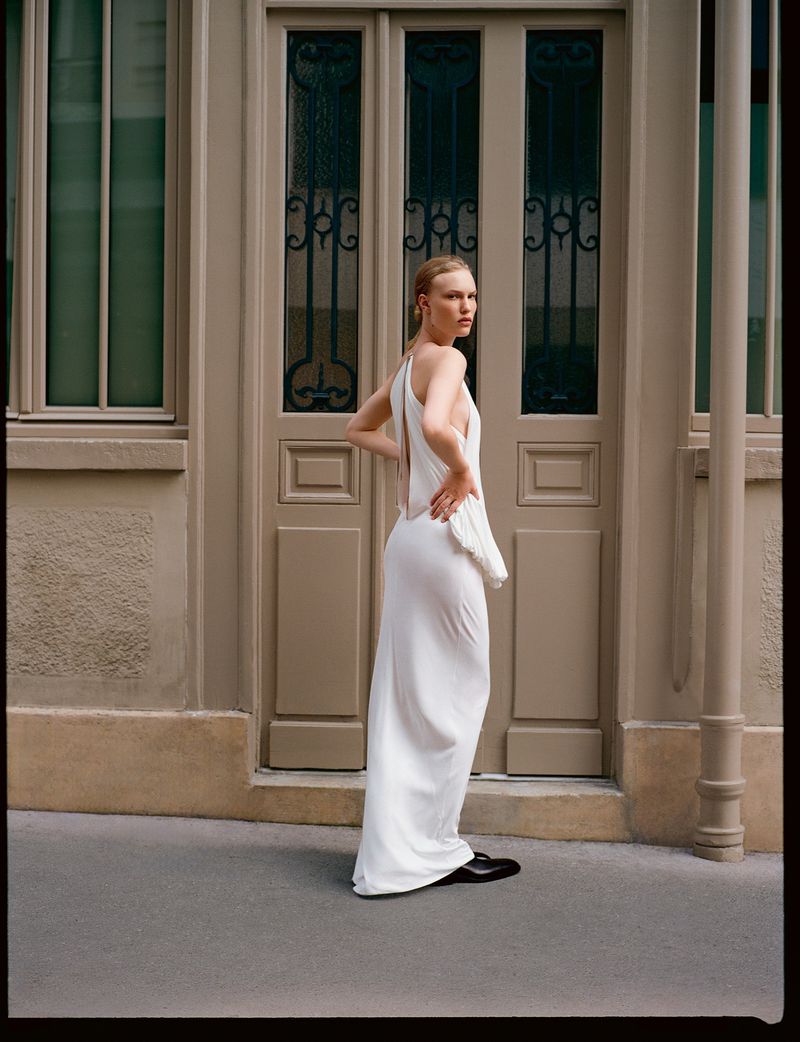 Elegant woman in a white gown poses in an outdoor setting, showcasing a modern fashion style against a neutral-toned building facade.