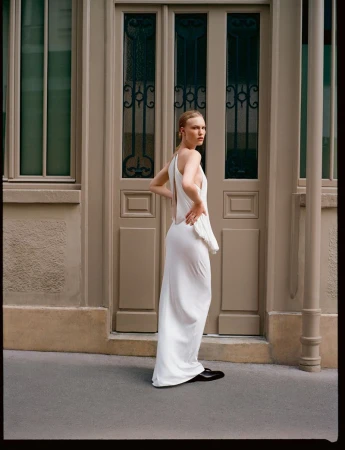 Elegant woman in a white gown poses in an outdoor setting, showcasing a modern fashion style against a neutral-toned building facade.