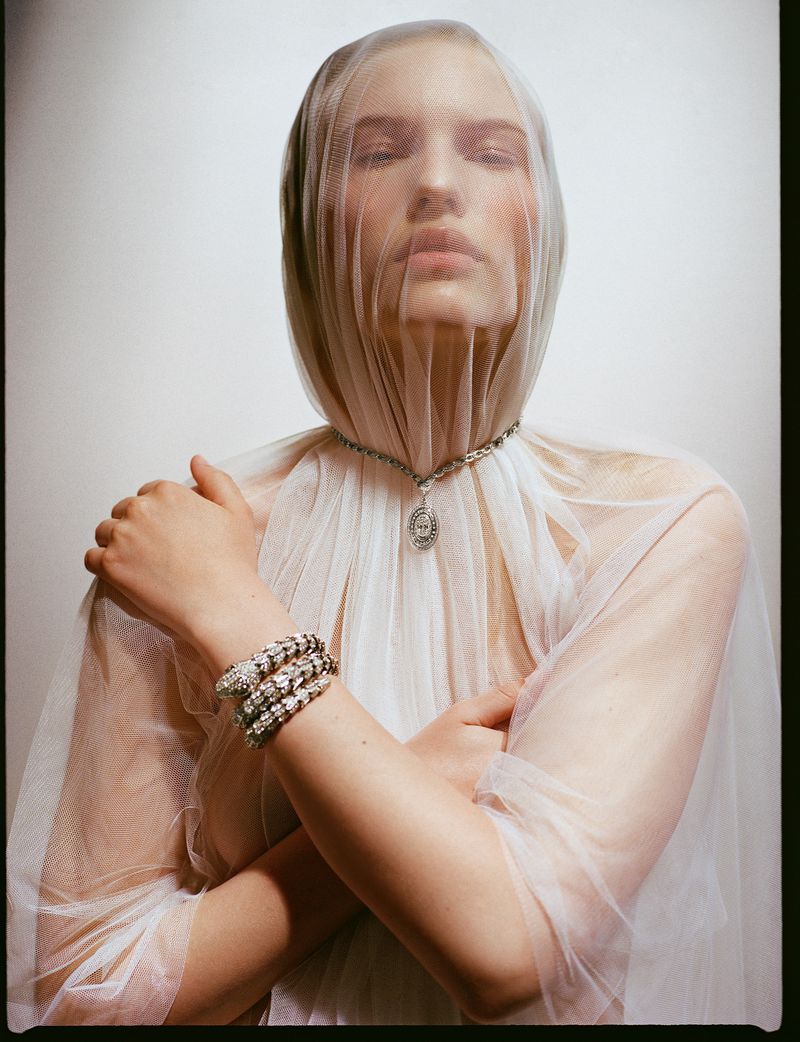 Close-up studio shot of a fair-skinned adult female model with folded arms, necklace, bracelets, and a sheer veil over her head.