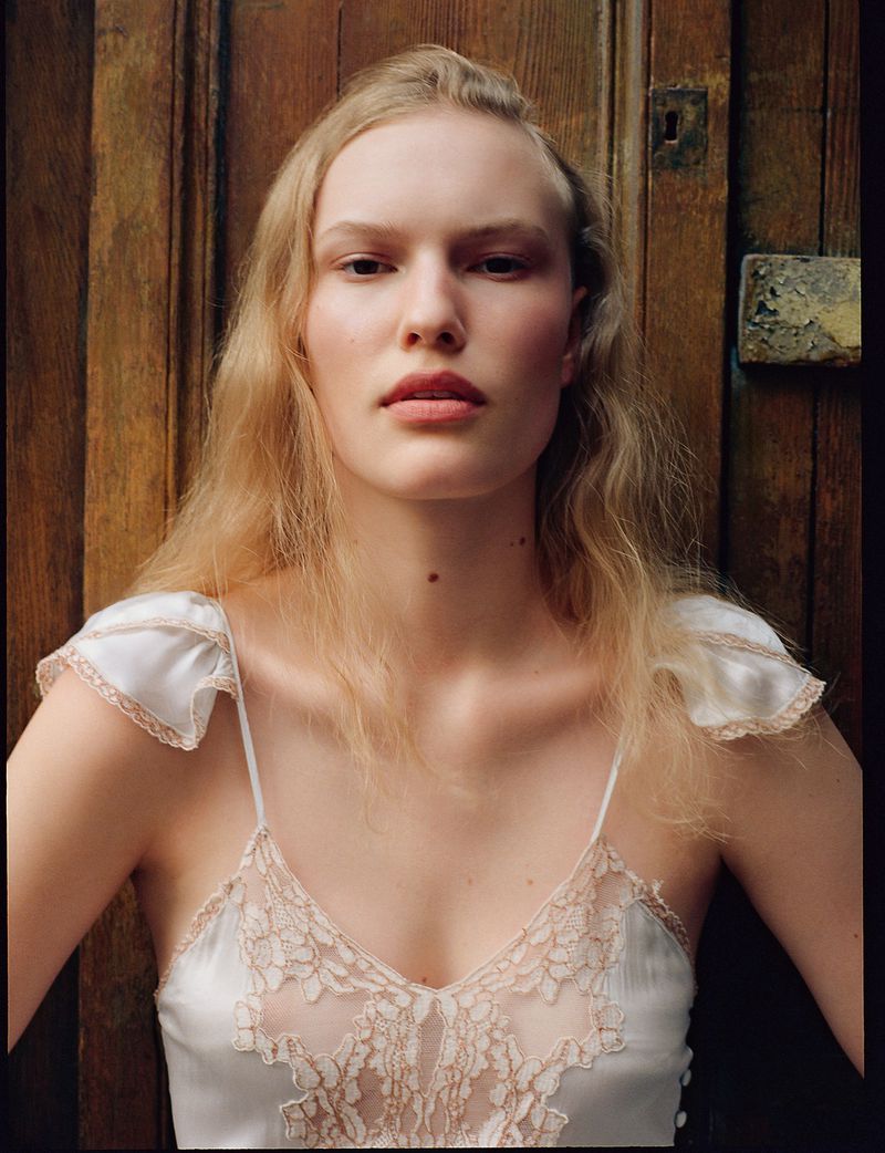 Portrait of a blond woman with fair skin, posing indoors with a wooden door background, wearing a white and lace top, medium shot.