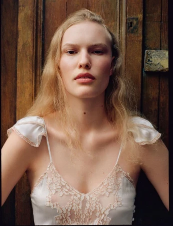 Portrait of a blond woman with fair skin, posing indoors with a wooden door background, wearing a white and lace top, medium shot.