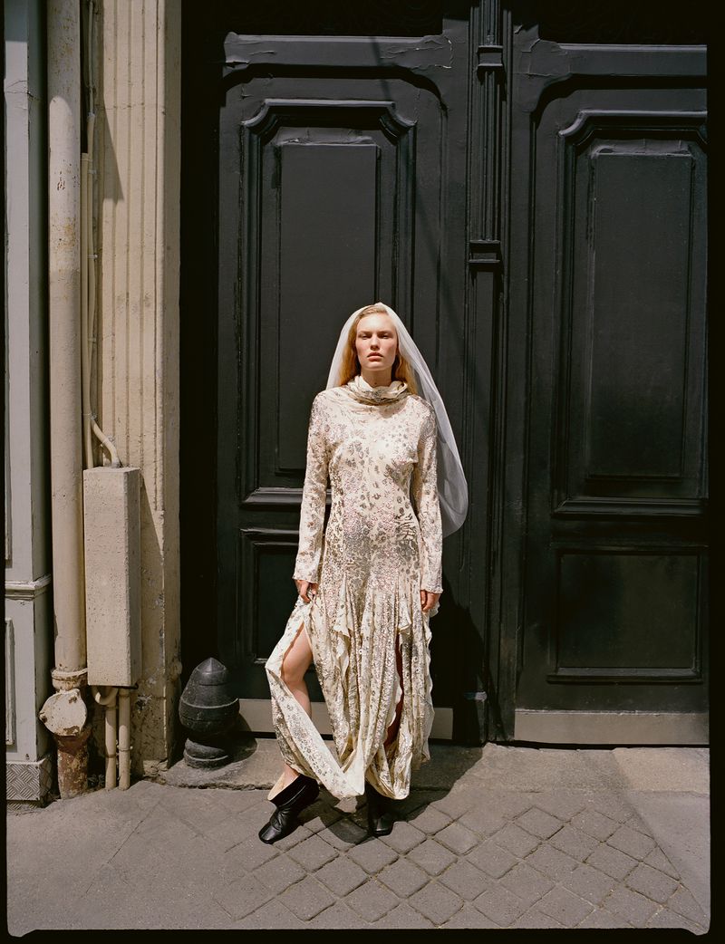 Woman in a long-sleeve patterned dress and veil poses against a dark door in an urban setting, creating a blend of fashion and location.
