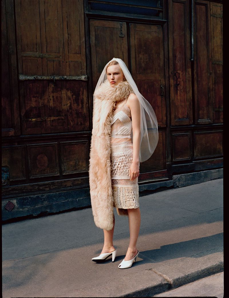 A blonde woman wearing a wedding dress, veil, fur stole, and white mules poses on a sidewalk in front of a dark wood door.