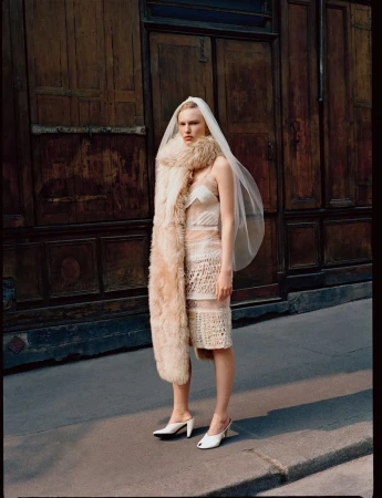 A blonde woman wearing a wedding dress, veil, fur stole, and white mules poses on a sidewalk in front of a dark wood door.