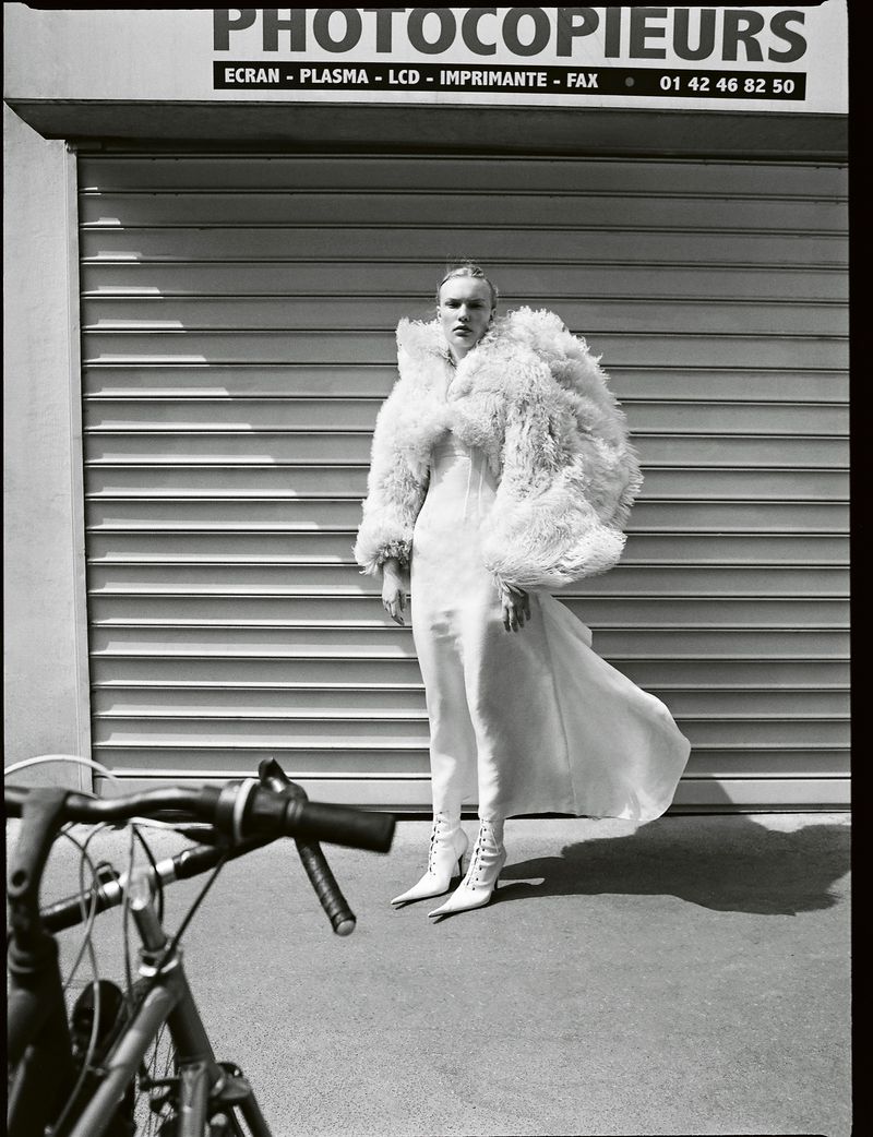Monochrome fashion shot of a woman in a long white dress and feathered coat standing on a city sidewalk with a bicycle in foreground.