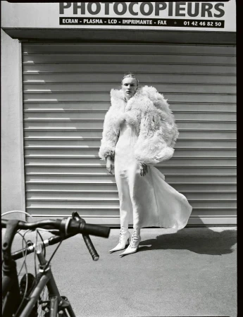 Monochrome fashion shot of a woman in a long white dress and feathered coat standing on a city sidewalk with a bicycle in foreground.