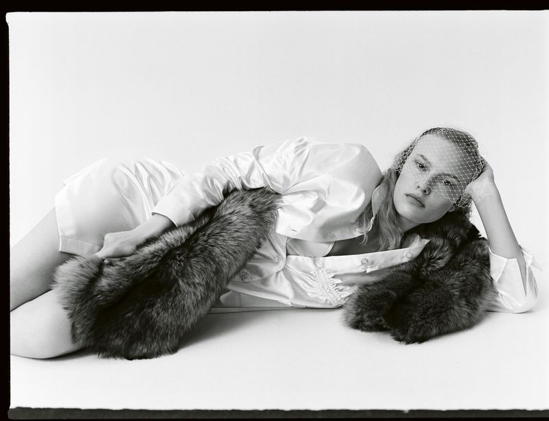 Monochrome fashion portrait of a woman lying down wearing a white dress, fur stole, and mesh veil against a white background.