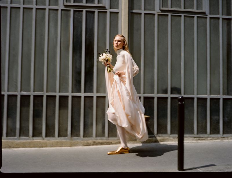 A woman in white dress and gold shoes walks on a city sidewalk holding flowers, with a gray metal gate in the background.