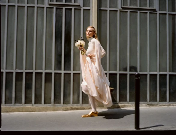 A woman in white dress and gold shoes walks on a city sidewalk holding flowers, with a gray metal gate in the background.