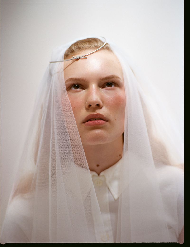 Close-up portrait of a fair-skinned woman with a sheer white veil and a silver headband, against a light background, looking up.