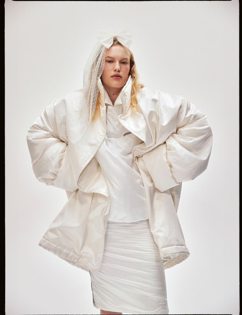 Fashion shot of a woman with blonde hair wearing a white padded coat, skirt, and a veil against a plain background in a studio.