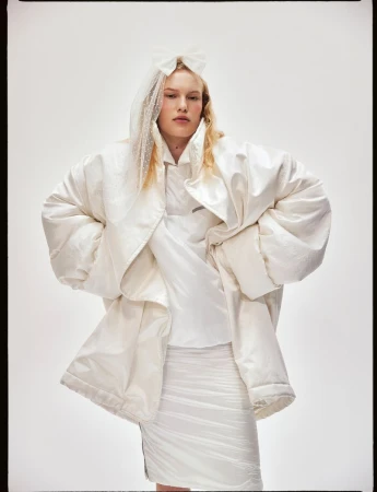 Fashion shot of a woman with blonde hair wearing a white padded coat, skirt, and a veil against a plain background in a studio.