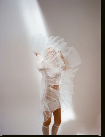 Woman poses in a sheer white ruffle dress against a neutral background with a beam of light, creating a soft, ethereal aesthetic.