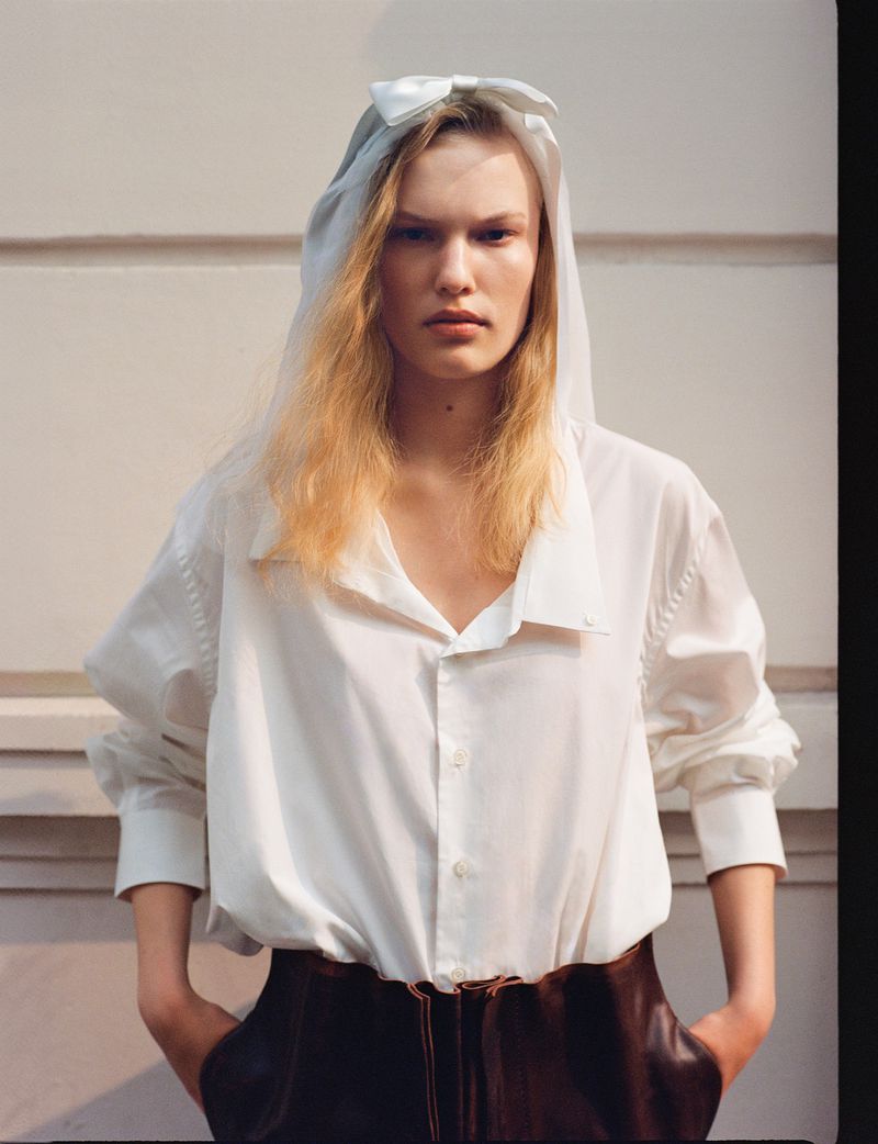 Fashion portrait of a fair-skinned young adult woman with light blonde hair wearing a white shirt and brown pants against a white wall.