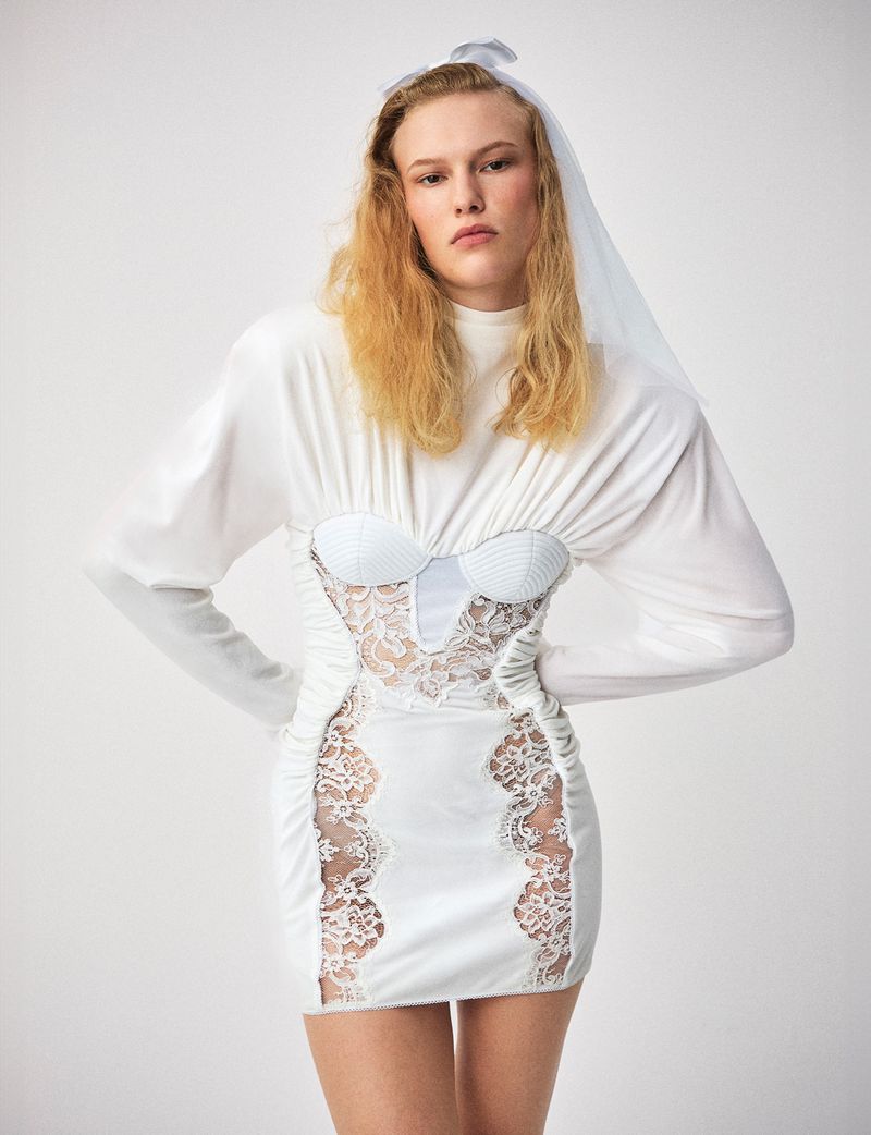 Woman with blonde hair wearing a white long-sleeve mini dress with lace on a white background in a studio setting.