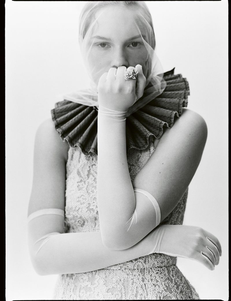 Monochrome portrait of a woman in elegant fashion, featuring a ruffled collar, veiled head, long gloves, and a prominent ring, in a studio setting.