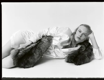 Model Astrid Østergaard reclining in a white satin shirt and birdcage veil with a fur stole against a white background.