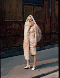 Model Astrid Østergaard poses outdoors wearing a white lace outfit, bridal veil, long fur stole, and white heels before a wooden doorway.