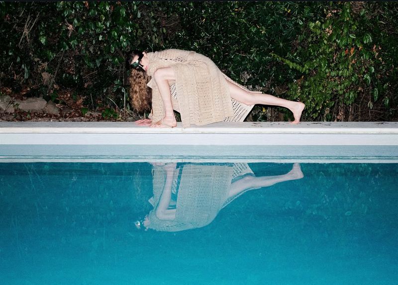 Woman in sheer knitwear poses at edge of pool with reflection in blue water, framed by green foliage in outdoor setting.