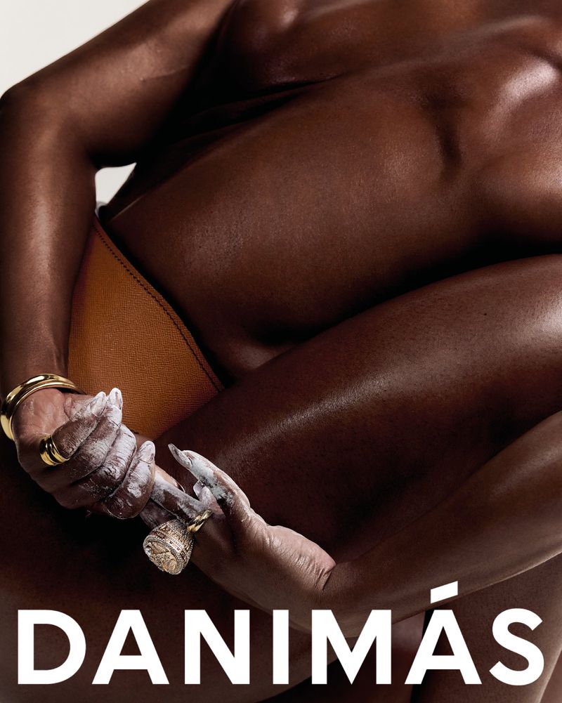 Close-up of two dark-skinned adult females embracing, one with white cosmetic cream on their hands, gold jewelry, and brown bikini bottoms.