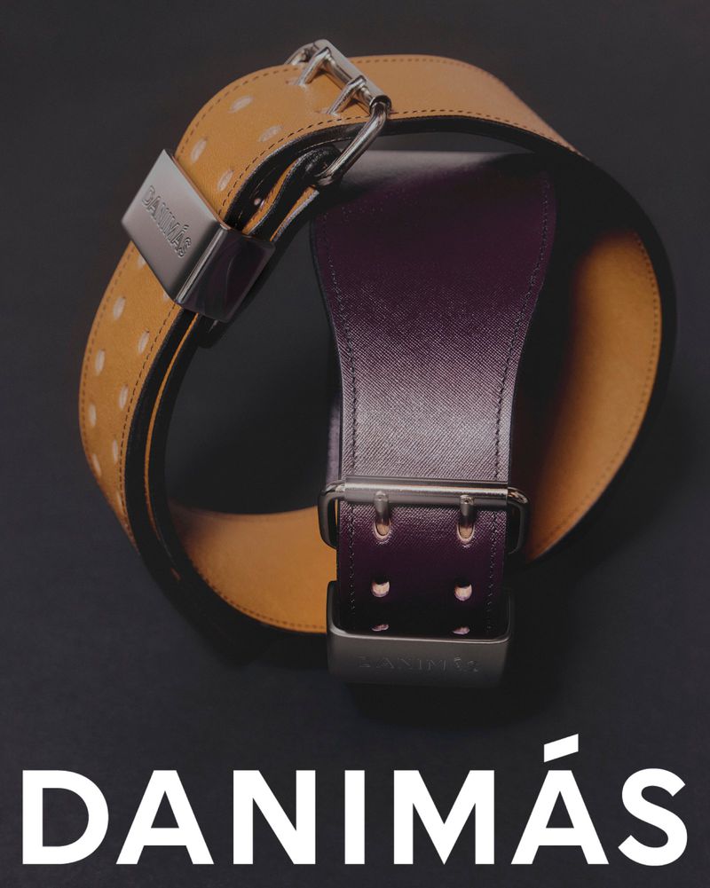 Two leather belts are displayed on a black surface, featuring silver buckles and a company logo at the bottom. Studio product shot.