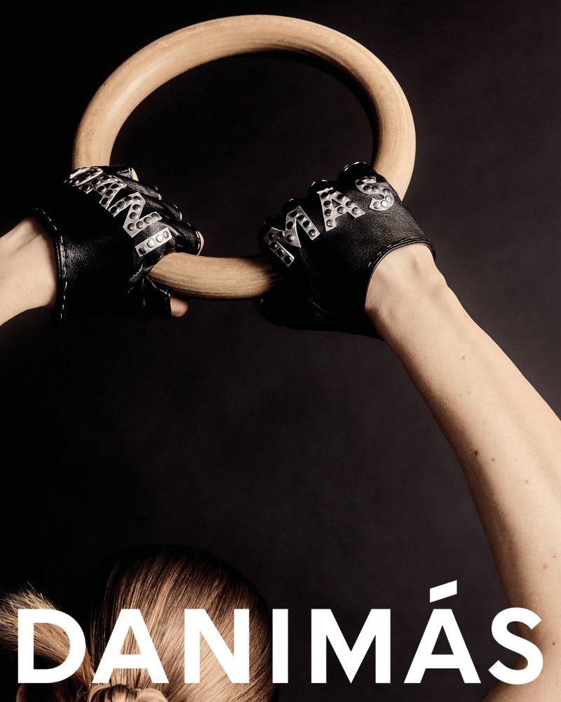 A woman's arms lift wooden gymnastic rings, hands in studded leather gloves against a stark black background with white text.