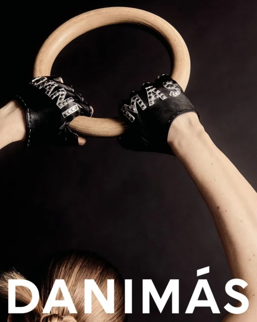 A woman's arms lift wooden gymnastic rings, hands in studded leather gloves against a stark black background with white text.