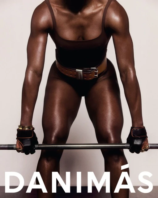 Athletic woman in a brown bodysuit prepares to lift a barbell, showcasing strength and fitness against a plain studio backdrop.