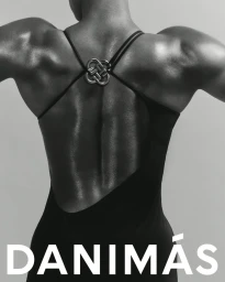 Monochrome rear-view portrait of a muscular person in a backless black dress with cross straps and metallic hardware, DANIMÁS typography below.