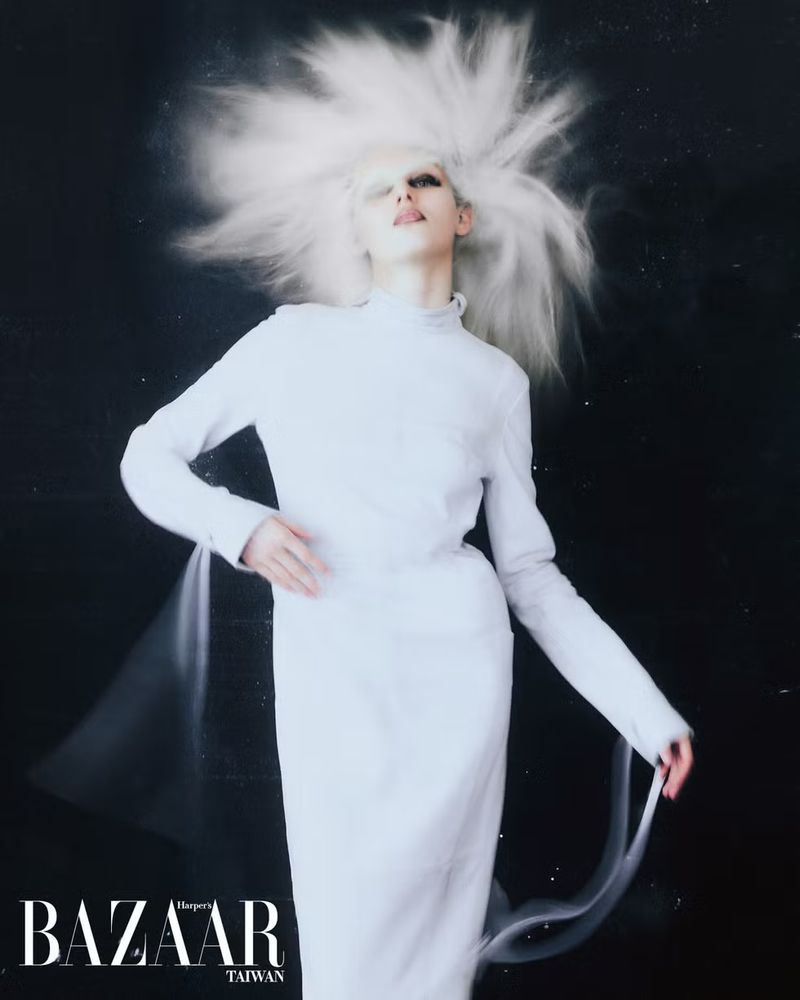 Fashion photo featuring a slender adult model with unique white hairstyle and makeup, wearing a white dress against a dark backdrop.
