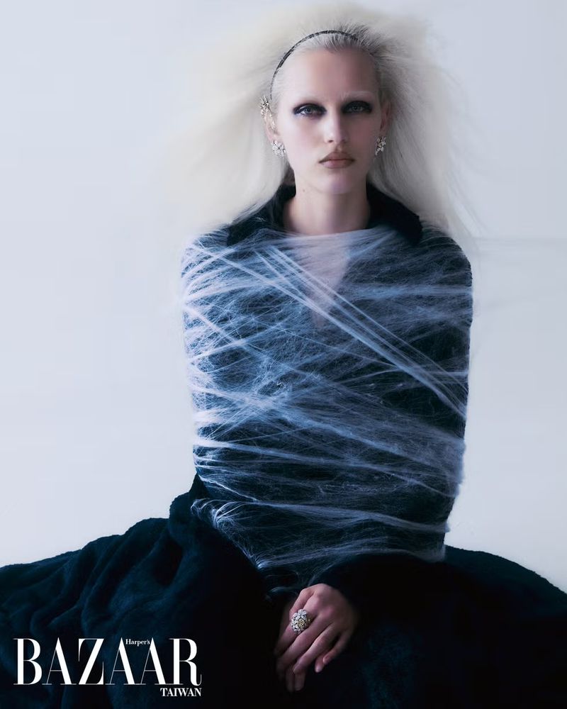 Fashion editorial portrait of a model wrapped in a spider web wearing black clothing with platinum hair and dramatic eye makeup against white.