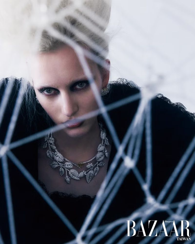 Fashion editorial shot of a fair-skinned woman with platinum hair wearing a leaf-patterned necklace, seen through a spiderweb-like pattern.