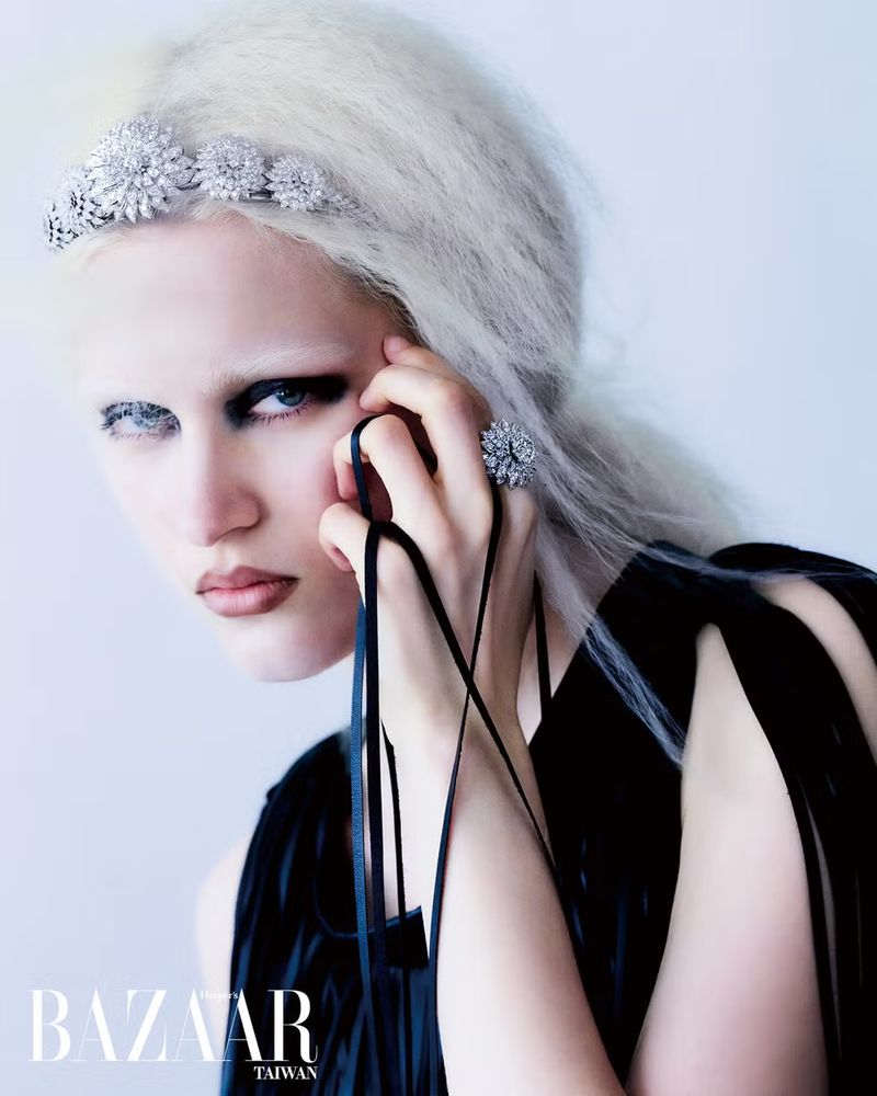 Close-up fashion shot of a model with white hair wearing an elaborate jeweled headband, statement ring, and black dress against a white background.