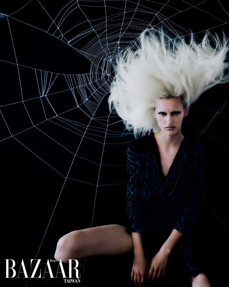 Editorial fashion shot of a woman with white hair and a dark dress sitting against a spider web background in a studio.