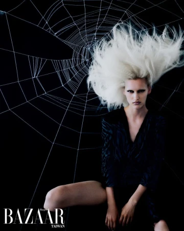 Editorial fashion shot of a woman with white hair and a dark dress sitting against a spider web background in a studio.