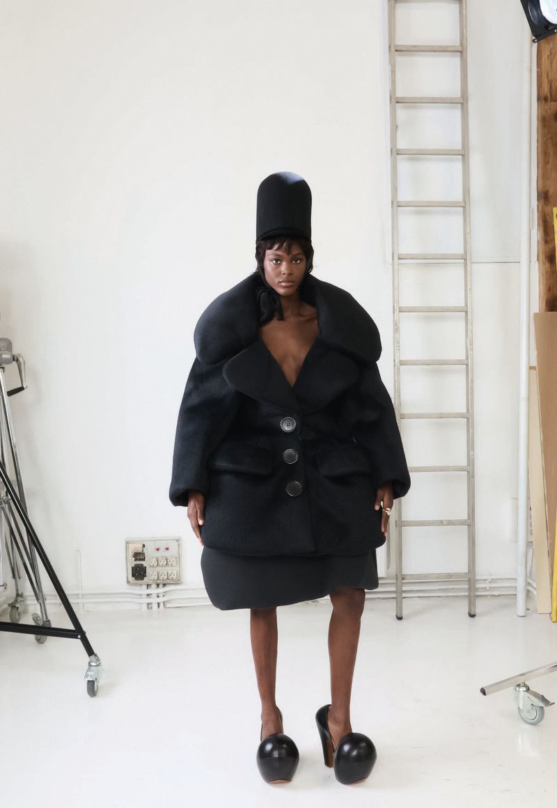 A fashion model poses in an oversized black coat, matching skirt and sculptural heels in a photo studio with natural light.