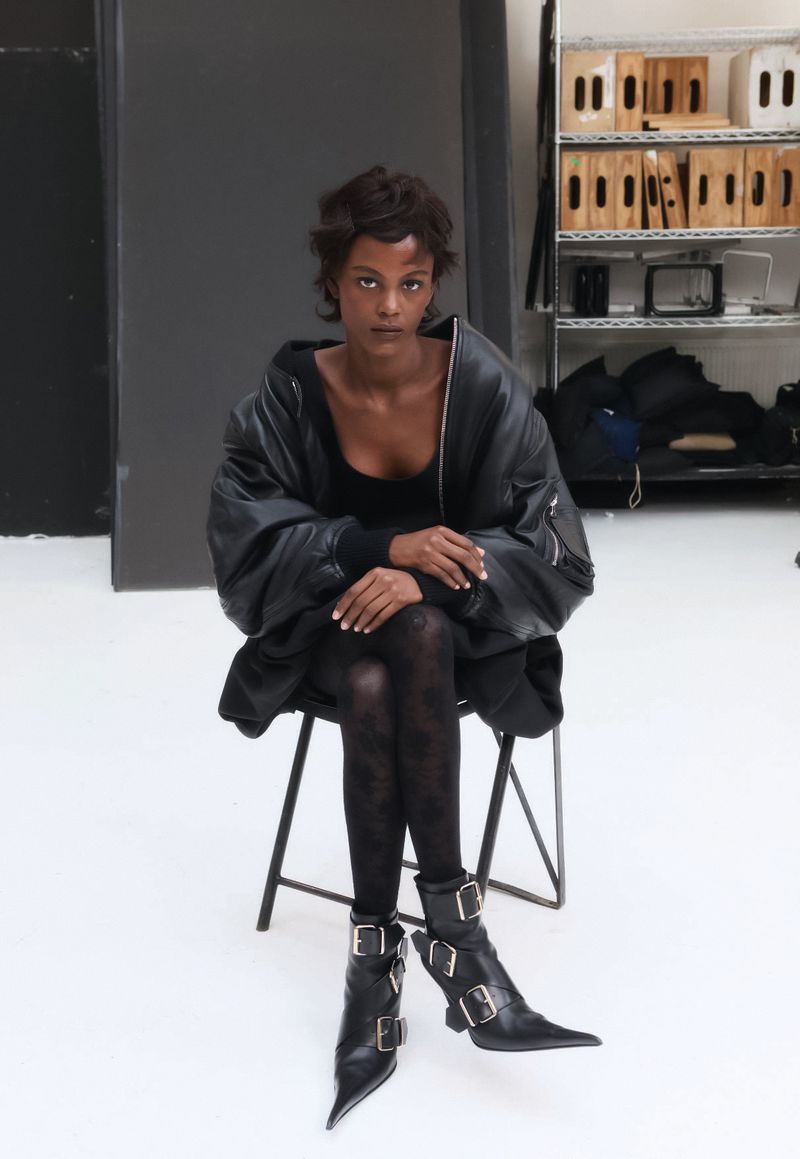 A model sits in a photo studio wearing a black bomber jacket, tights, and buckle boots in a fashion photography setting.
