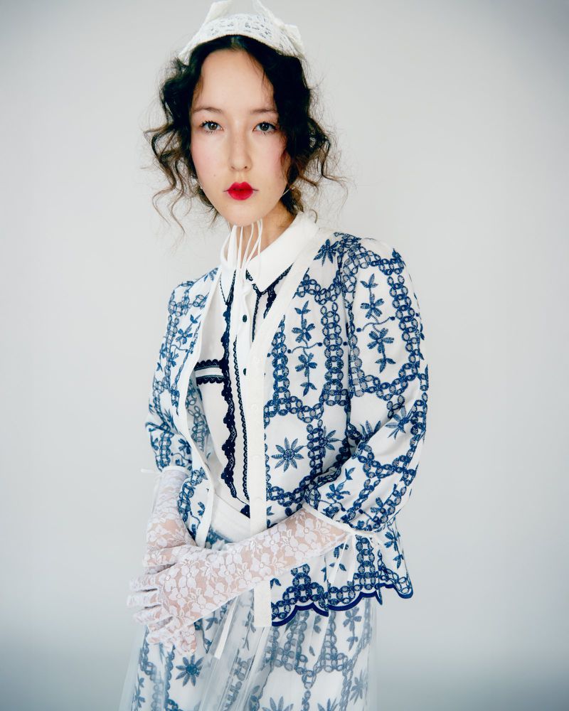 Woman poses in white and blue embroidered jacket, matching skirt, and lace gloves against a white backdrop in a studio fashion shot.