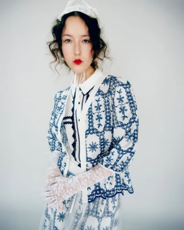 Woman poses in white and blue embroidered jacket, matching skirt, and lace gloves against a white backdrop in a studio fashion shot.