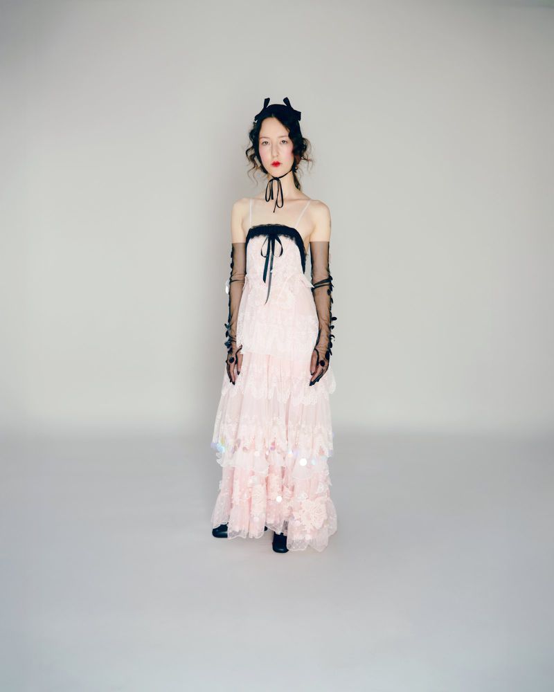 Model standing in a tiered pale pink tulle dress and sheer black opera gloves against a studio background for Bora Aksu S/S 26.