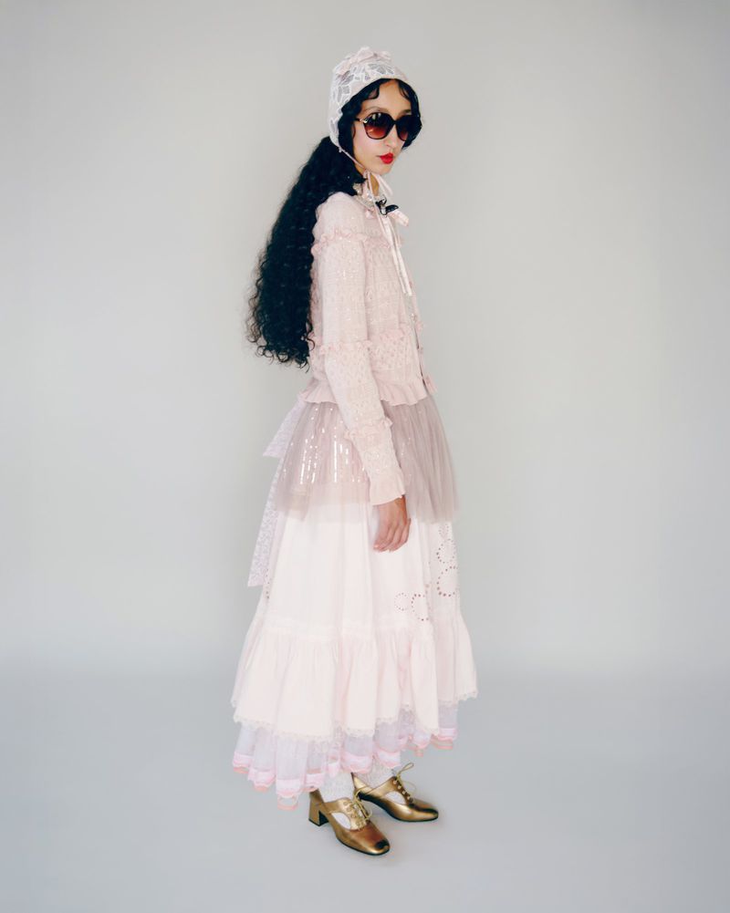 Full shot of a woman wearing sunglasses, a lace bonnet, a cardigan, and a layered tulle skirt. She also wears shiny gold shoes.
