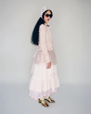 Full shot of a woman wearing sunglasses, a lace bonnet, a cardigan, and a layered tulle skirt. She also wears shiny gold shoes.