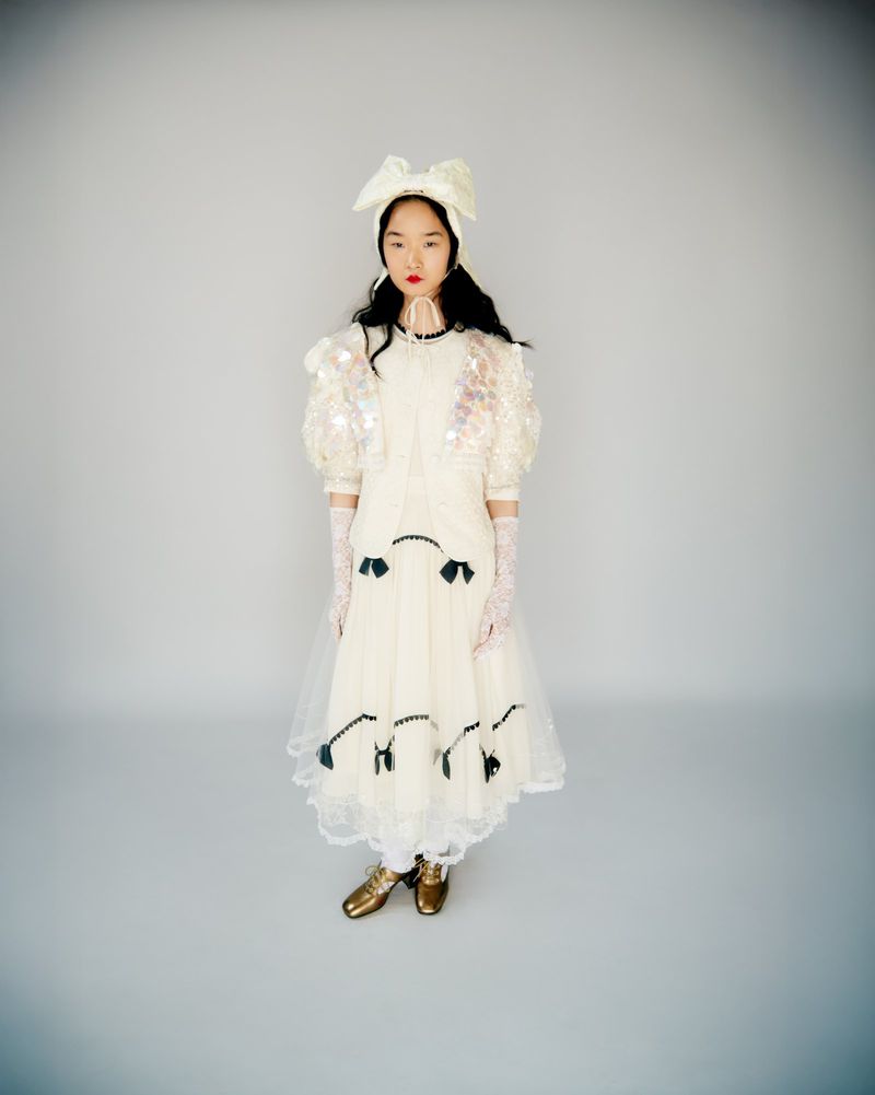 Young Asian woman poses indoors wearing an ornate white dress, gold shoes, gloves, and a bow headband against a neutral background.