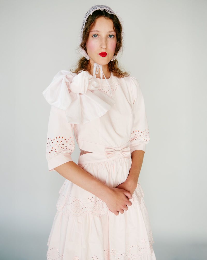 Young woman in a light pink dress with lace bonnet poses in front of a white background, accentuating the dress's design.