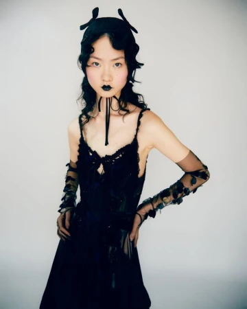 A young Asian woman poses in a black dress and headpiece against a light background, showcasing fashion and beauty with unique style.
